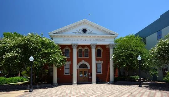 Red-brick Carnegie public library building with white columns, arched windows, and a triangular pediment, surrounded by trees on a sunny day.