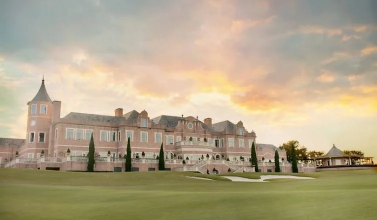 Large, elegant mansion with a tower by a well-manicured golf green under a colorful sunset sky.