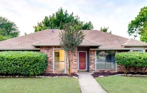 Single-story brick house with a red front door, large front windows, trimmed bushes, and a small tree, set against a backdrop of green trees and lawn.